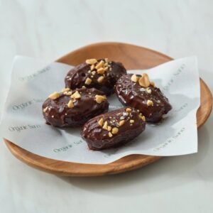 Four Organic Sano Snicker Date Bites, chocolate-coated dates with peanuts and sea salt on a wooden plate